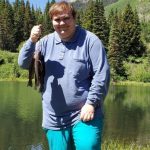 William, smiling and holding up a fish on a line, in front of a mountain lake