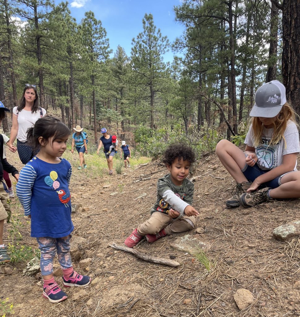 Three young children in foreground, one is pointing excitedly to something on the ground while the other two look on. Four adults hiking up hill behind them, one is carrying another small child. Lots of ponderosa trees and blue sky in background.